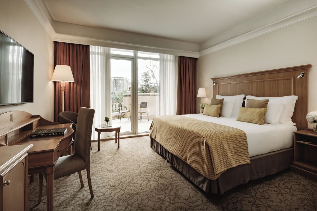 A hotel room with a large bed, a desk with a computer, a chair, a TV, and a view of the outdoors through a sliding door.