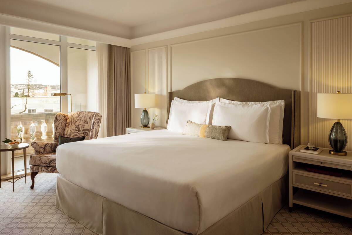 A large bed with a headboard, two bedside tables, a chair, and a view of the city through large windows.
