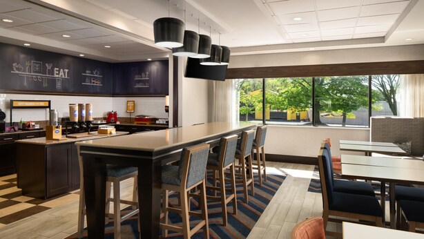 A modern hotel kitchen with a bar area, seating for guests, and large windows offering a view of the outdoors. - Hampton Inn Chambersburg (Chambersburg)