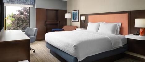 A hotel room with a large bed, a desk, a chair, and a view of trees outside the window.