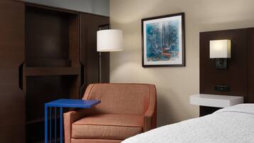 Habitación de hotel con cama, una mesita auxiliar azul, un sillón rojo, un cuadro enmarcado de un bosque, un lavamanos fijado a la pared y dos apliques.