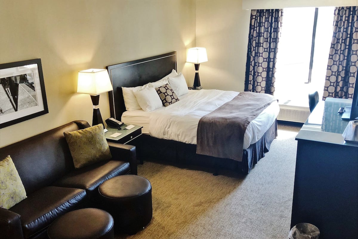A hotel room with a bed, two bedside lamps, a desk with a computer, a chair, and a small round ottoman.