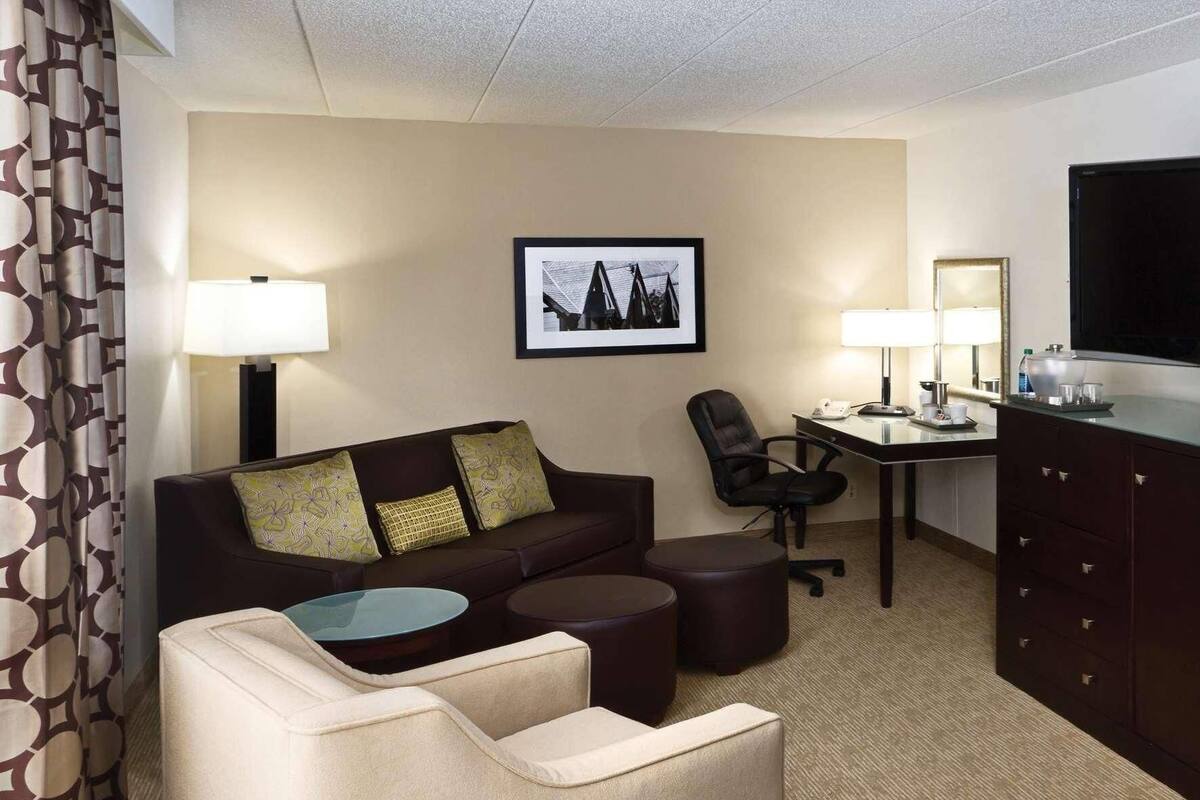 A hotel room with a sofa, two armchairs, a small round table, a desk with a chair, a television, and a minibar.