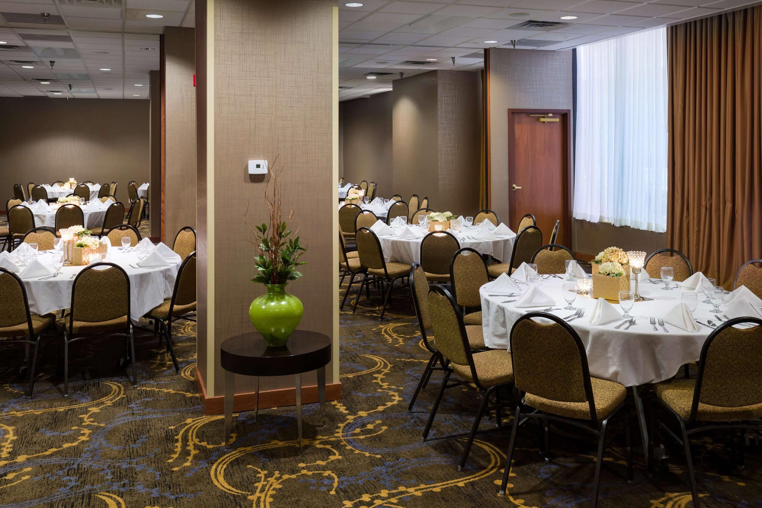 Une salle de conférence avec plusieurs tables dressées pour un repas, équipée de chaises, de tables et d’un vase décoratif.