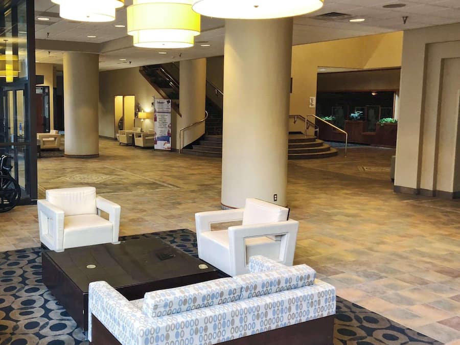 A lobby with a reception desk, white armchairs, a patterned sofa, and a staircase in the background.