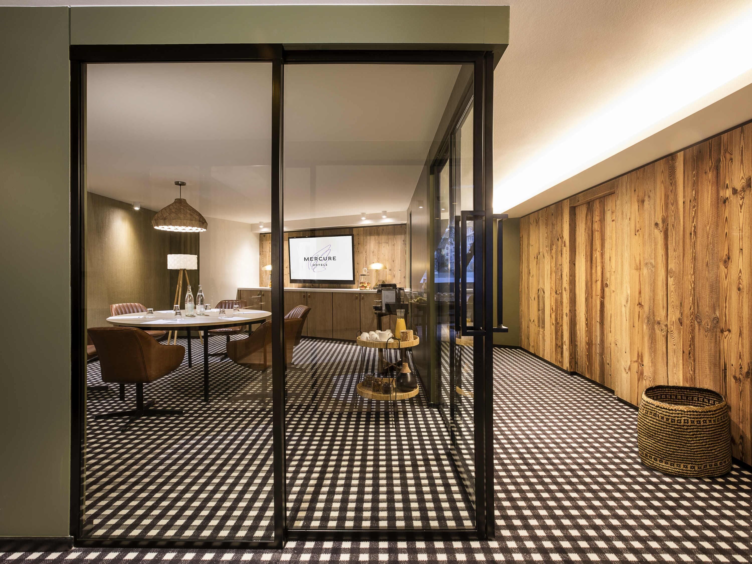 A hotel room with a checkered floor, a dining area with a round table, chairs, and a television screen displaying 'Mercure Hotels'.