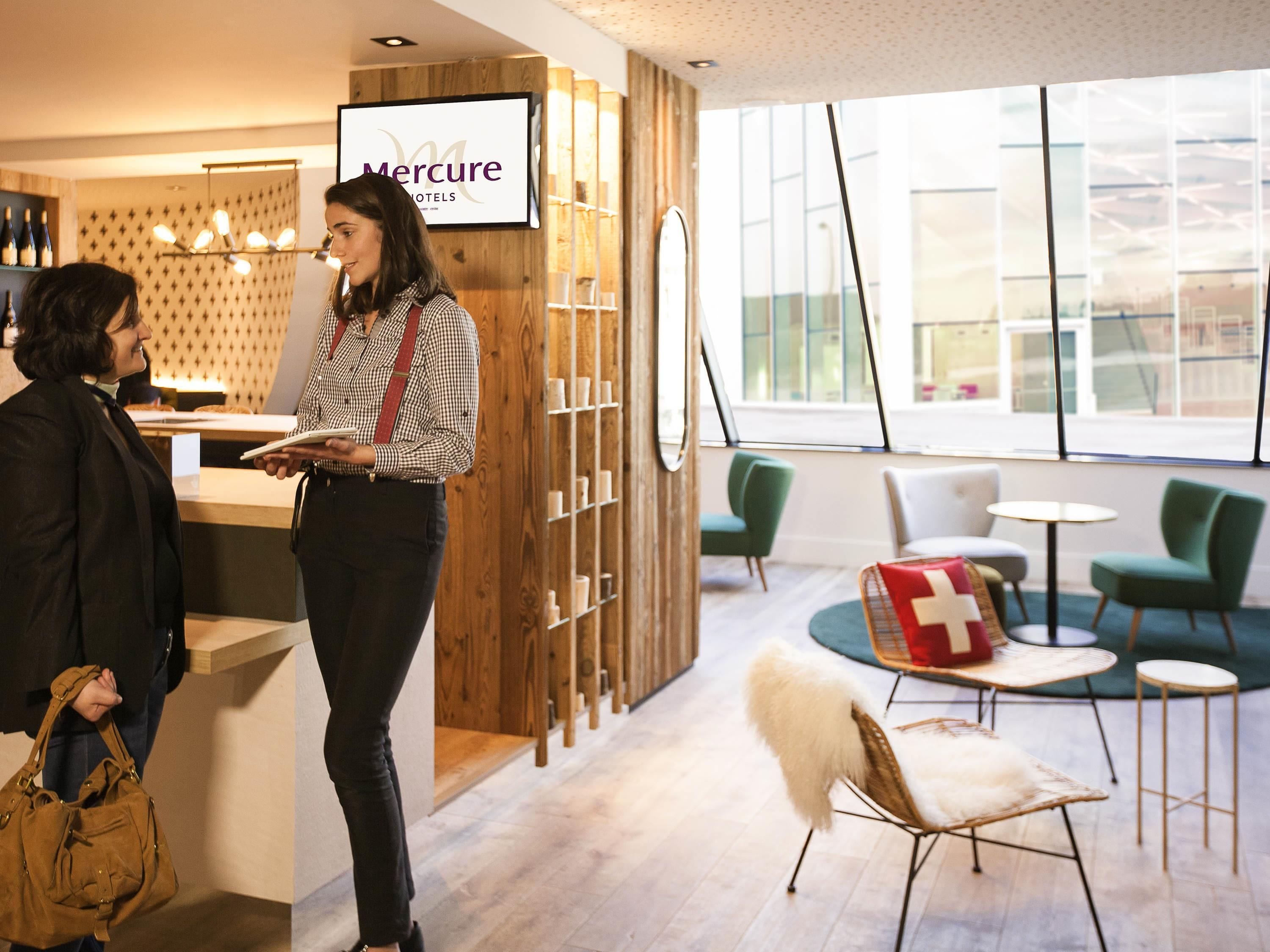 Zona de recepción de hotel con un mostrador de madera, un cartel de Mercure Hotels y vista del interior a través de amplios ventanales.