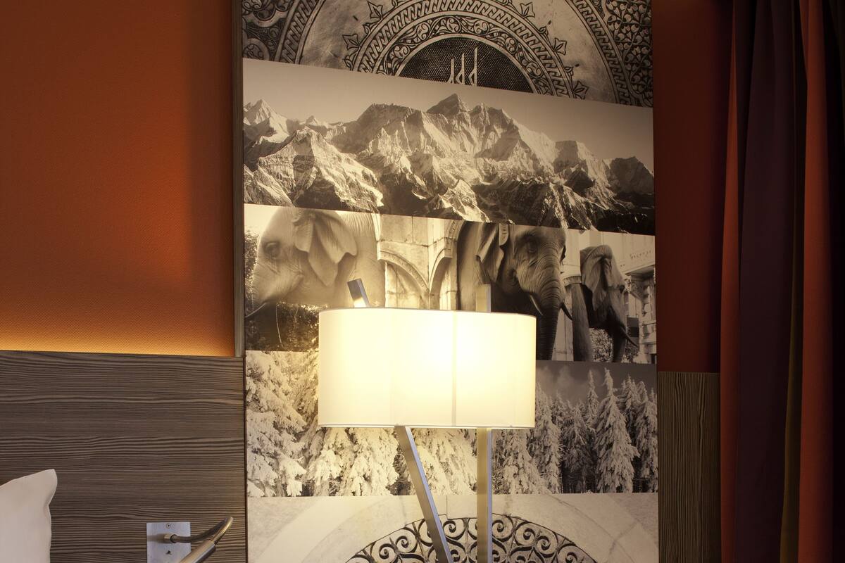 Habitación de hotel con cama, mesita de noche con lámpara, pared con un panel decorativo grande y un sillón de cuero marrón.