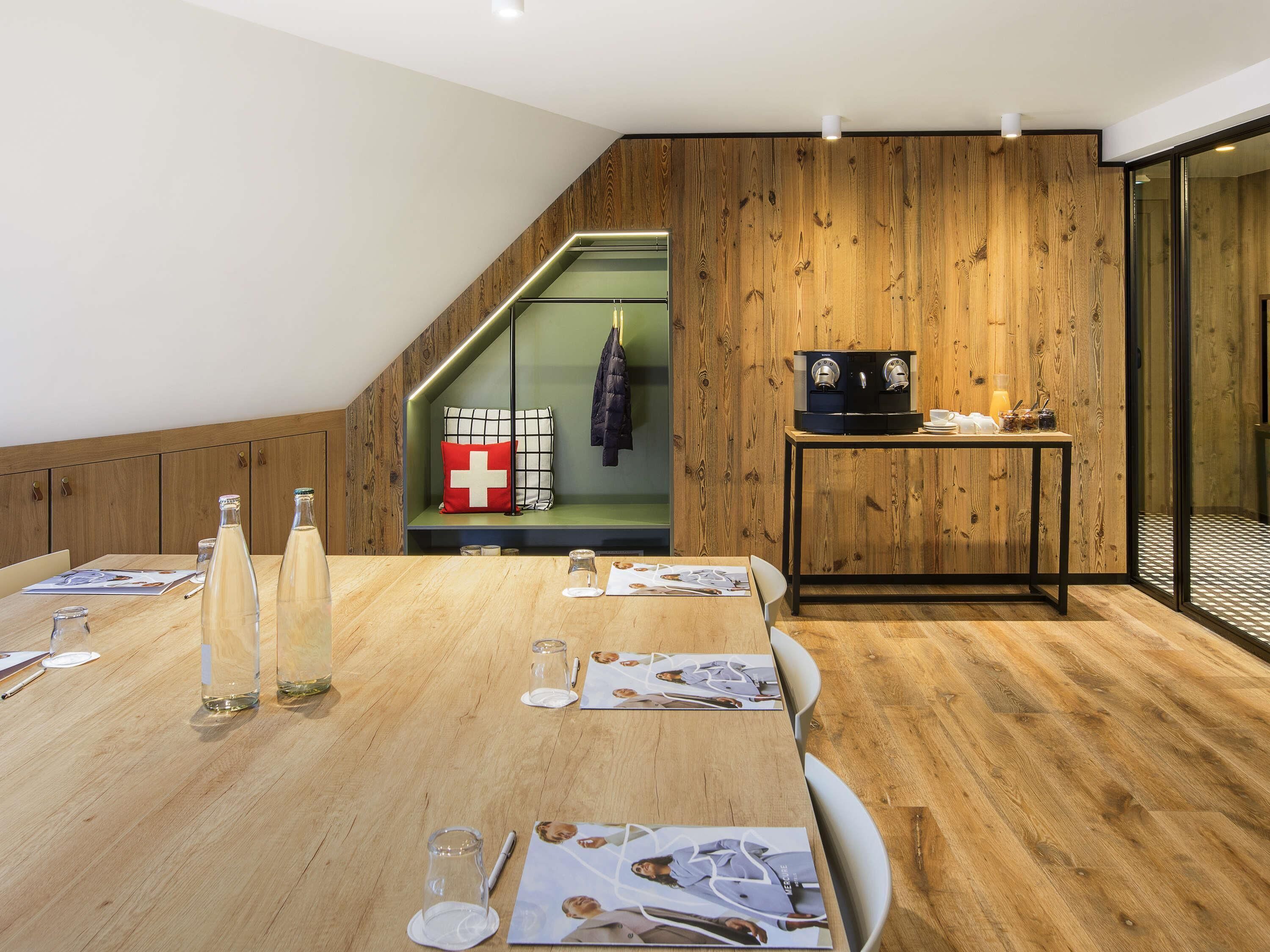 Una sala de reuniones moderna con suelo de madera, una mesa larga con revistas, una máquina de café y un perchero.