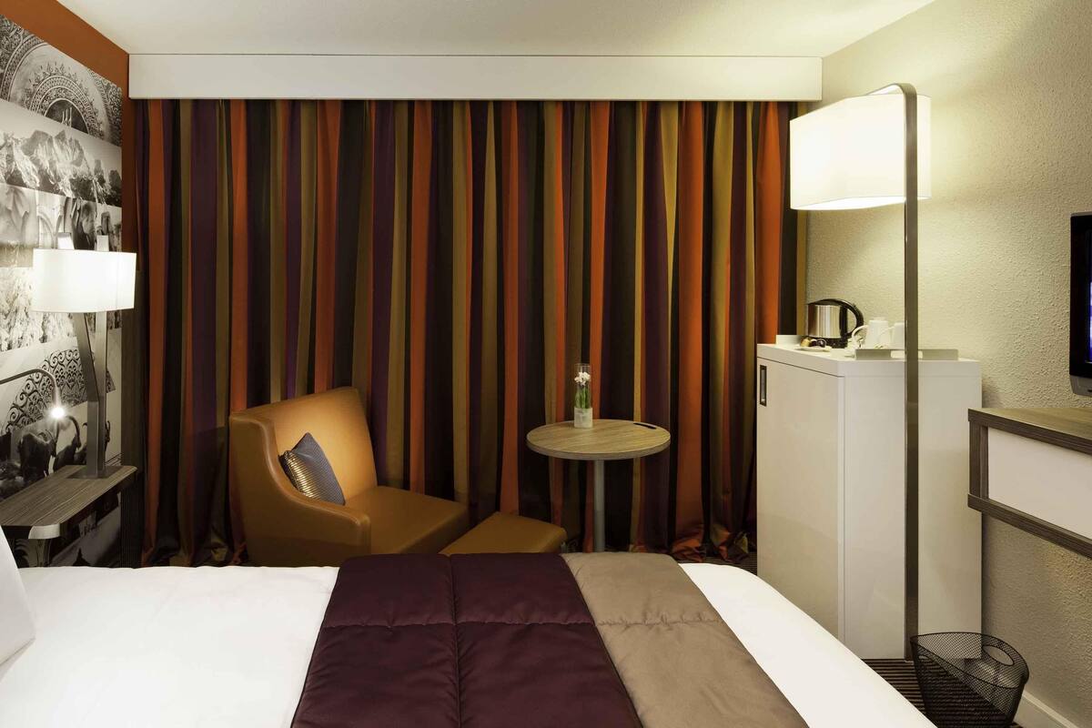 Habitación de hotel con cama, una silla, una mesita con un florero, un escritorio con lámpara y un refrigerador.