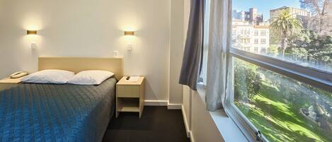 A hotel room with a bed, bedside lamps, a nightstand, a window with curtains, and a view of buildings and trees.