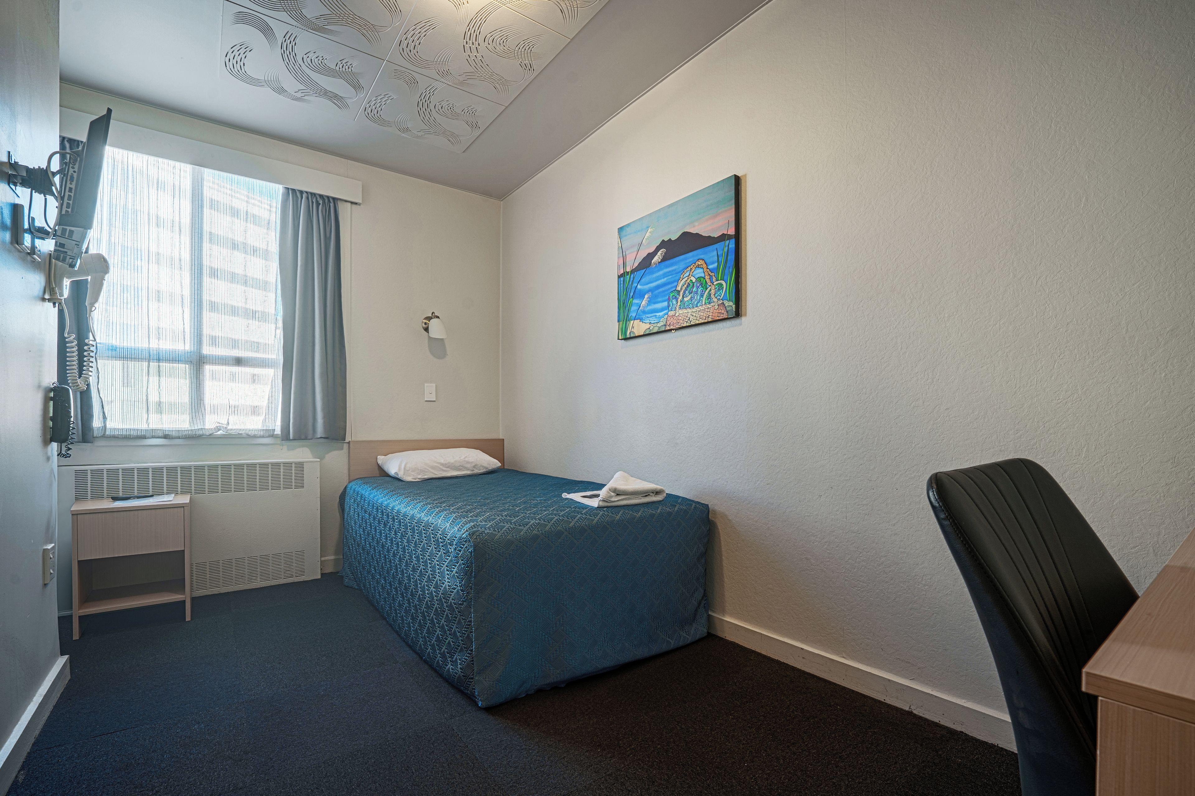 A hotel room with a single bed, a desk, a chair, a radiator, and a painting on the wall.