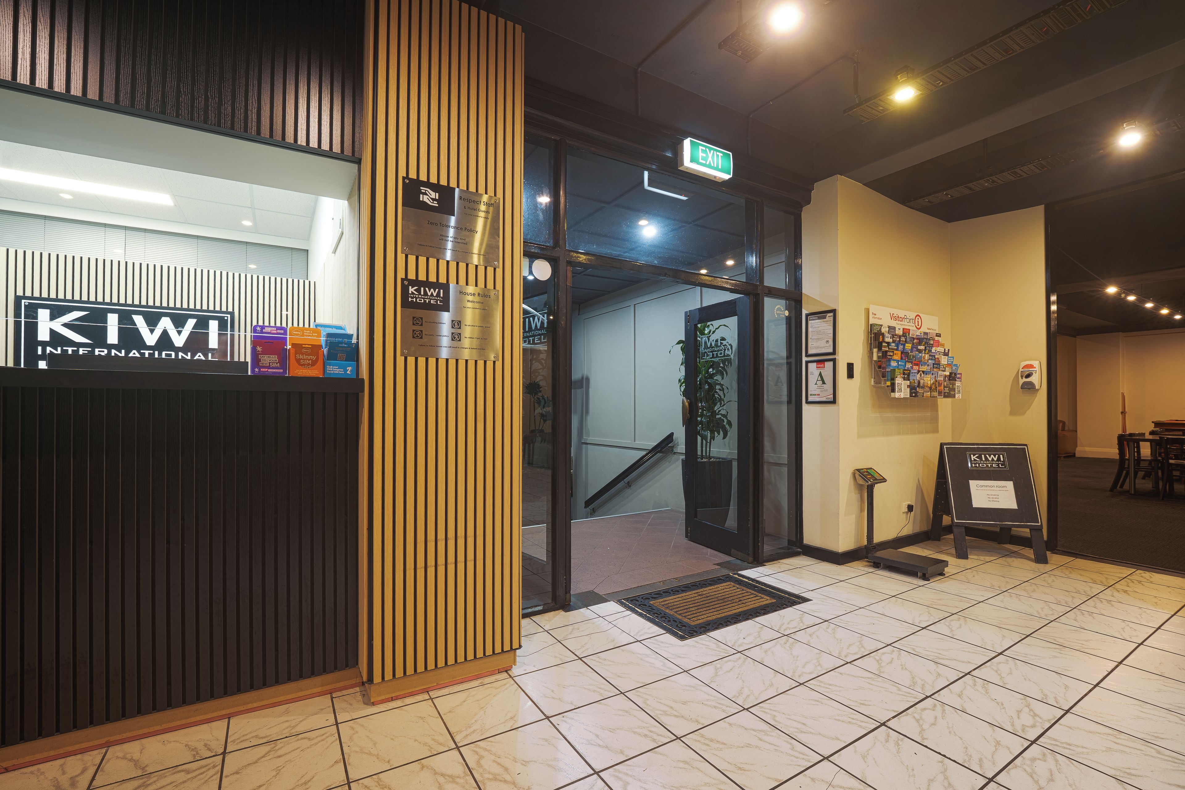Ingresso di un hotel con pannelli in legno, indicazione di uscita e reception con brochure informative.