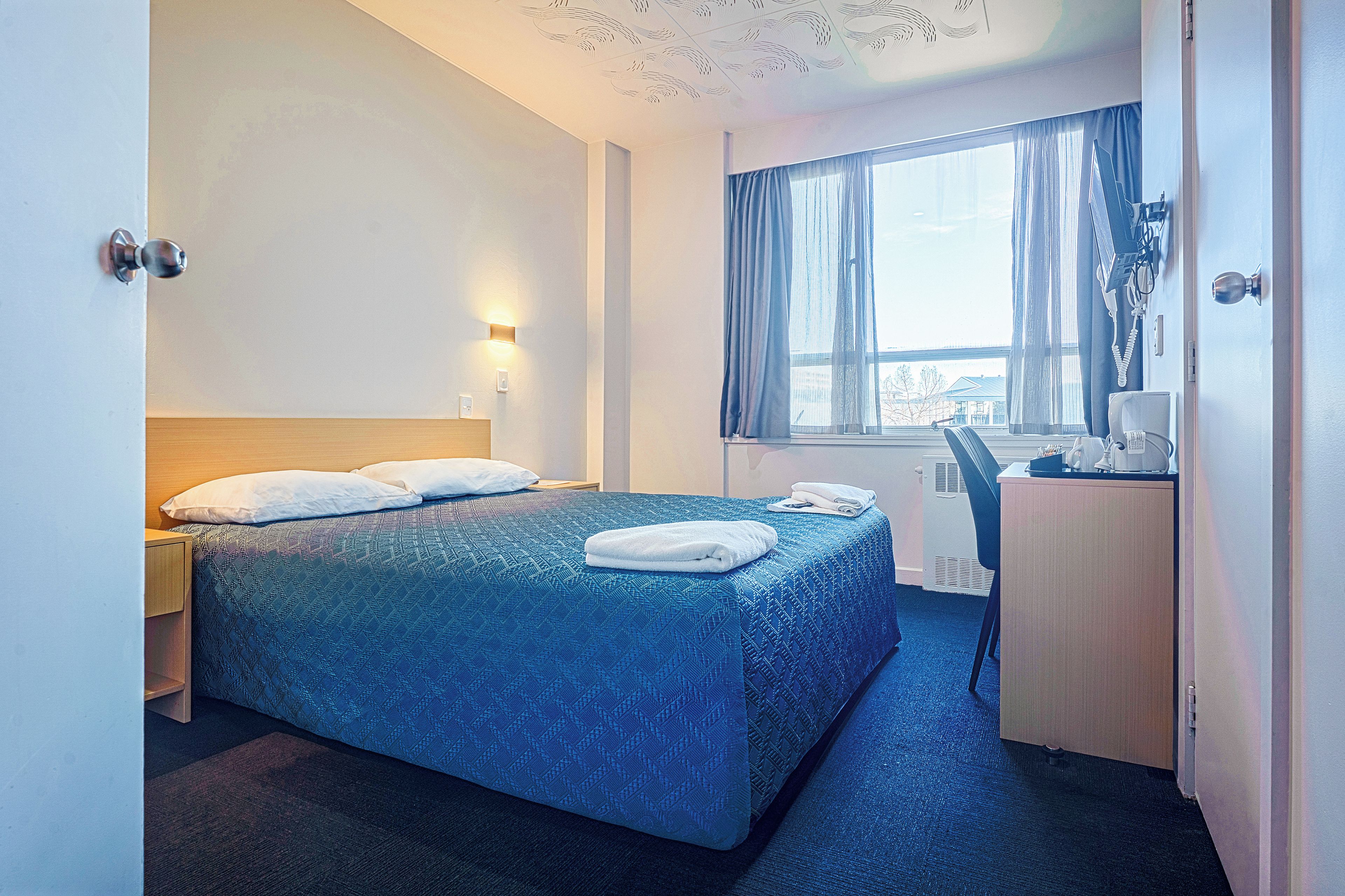 Habitación de hotel con cama, escritorio, silla, ventana con cortinas y una lámpara fijada a la pared.