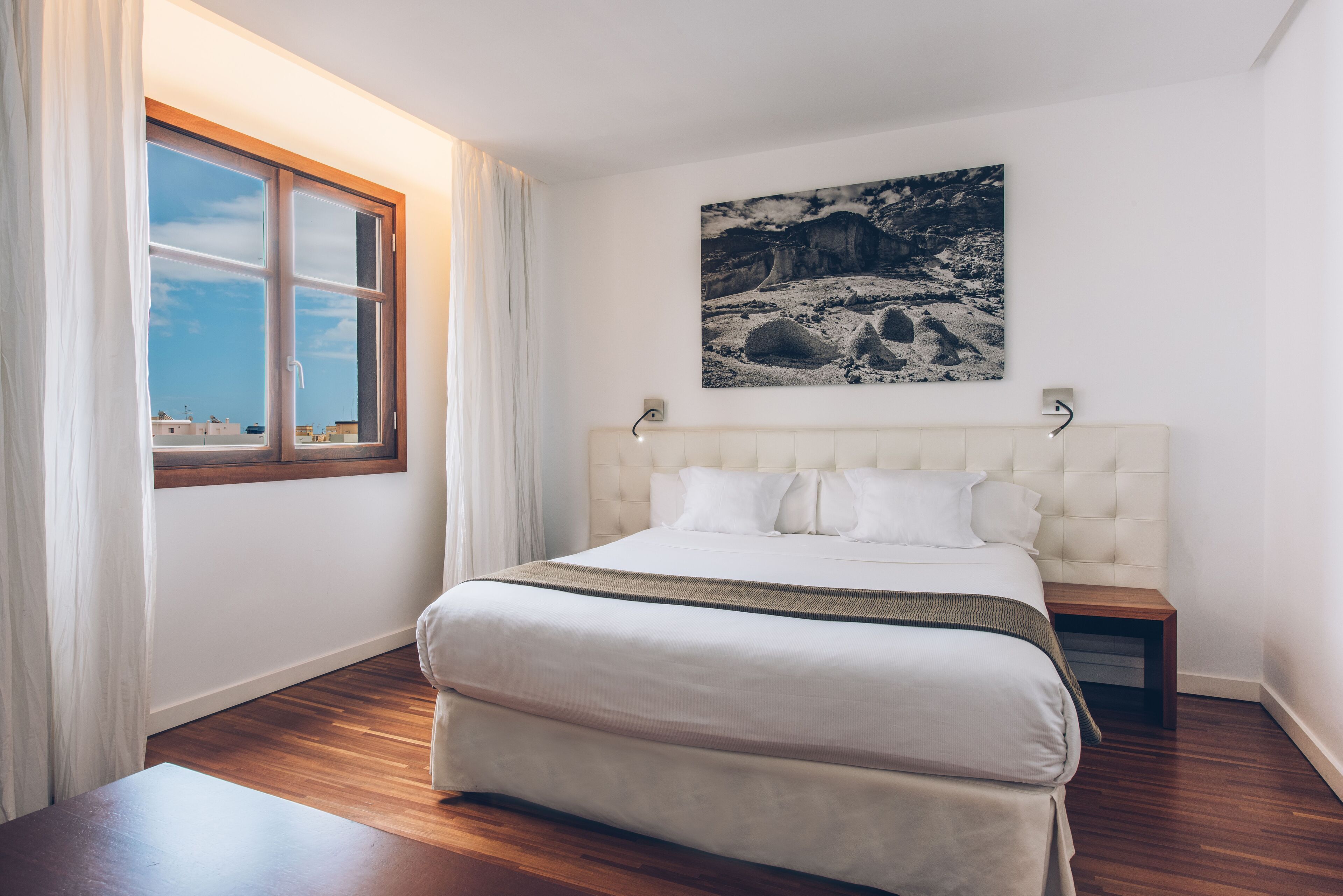Un dormitorio con una cama grande, una ventana con cortinas, una mesita de noche de madera y un cuadro colgado en la pared.