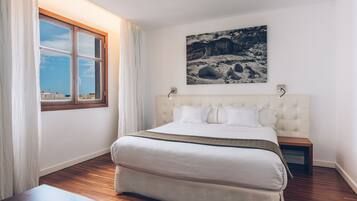 A bedroom with a large bed, a window with curtains, a wooden nightstand, and a wall-mounted picture.