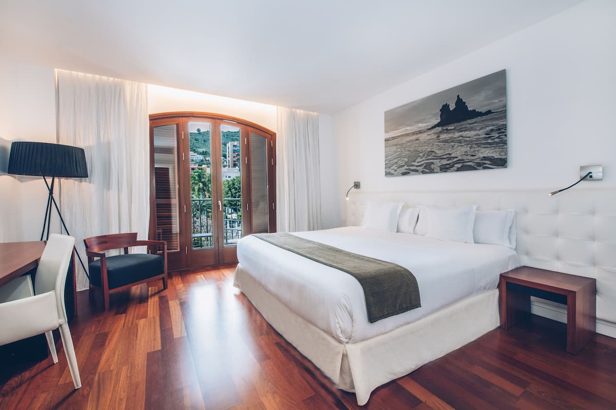 A hotel room with a large bed, a desk with a chair, a small table, and a view of the outdoors through a glass door.