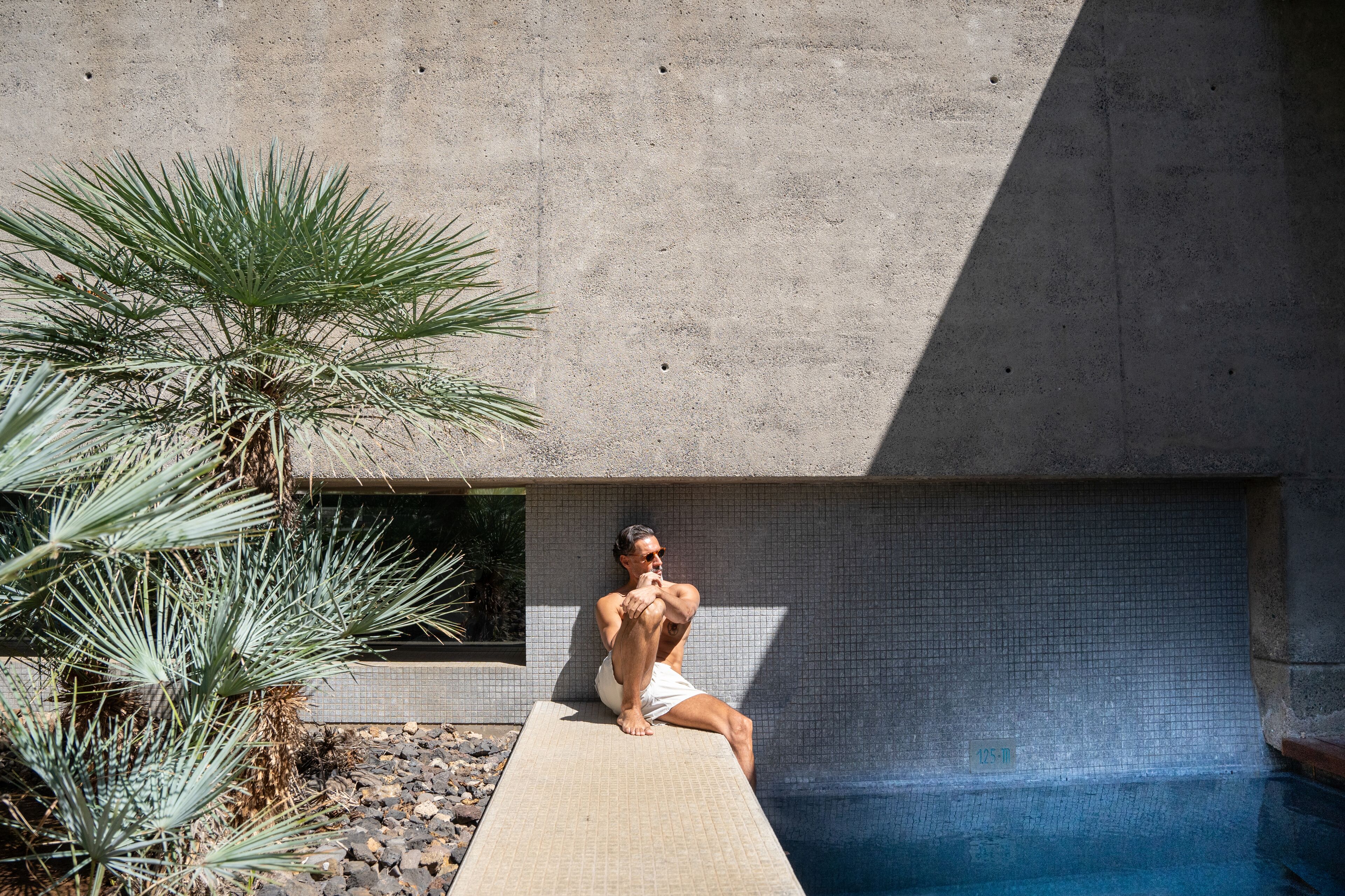 A man sitting on a poolside ledge with a palm tree nearby.