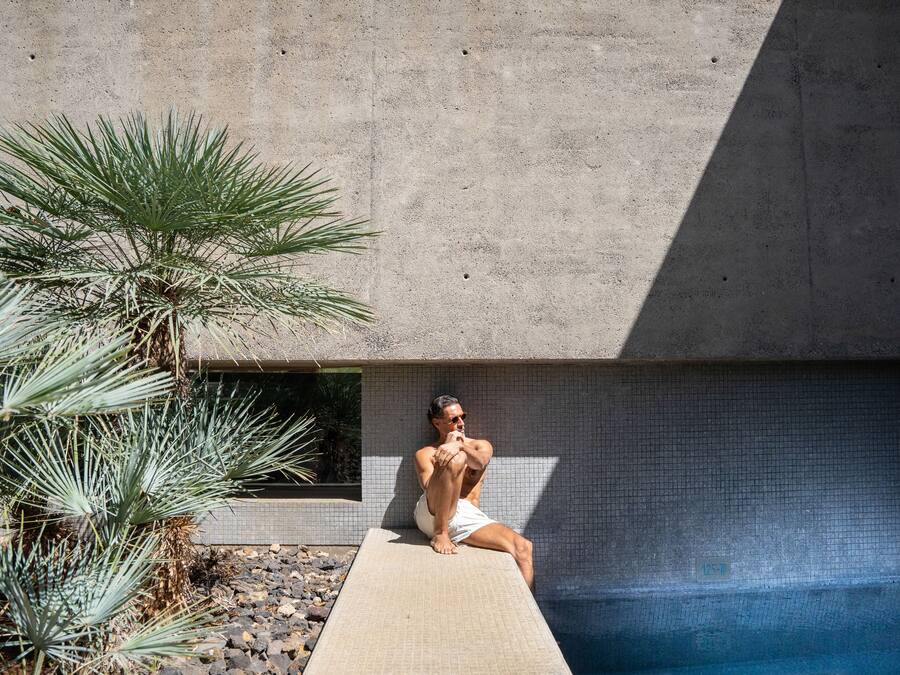 A man sitting on a poolside ledge with a palm tree nearby.