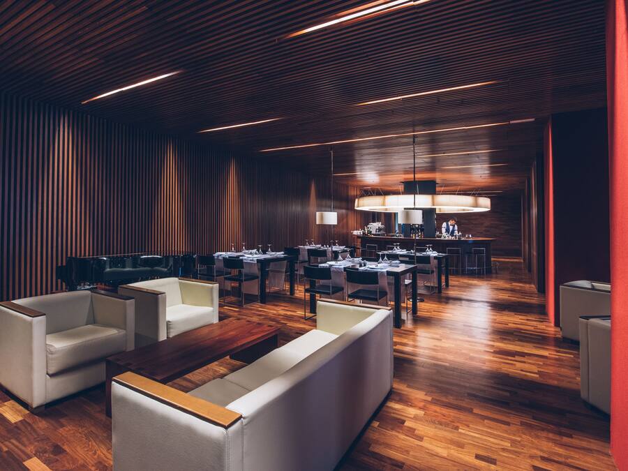 A modern restaurant with wooden flooring, white sofas, and a central dining area with tables and chairs.