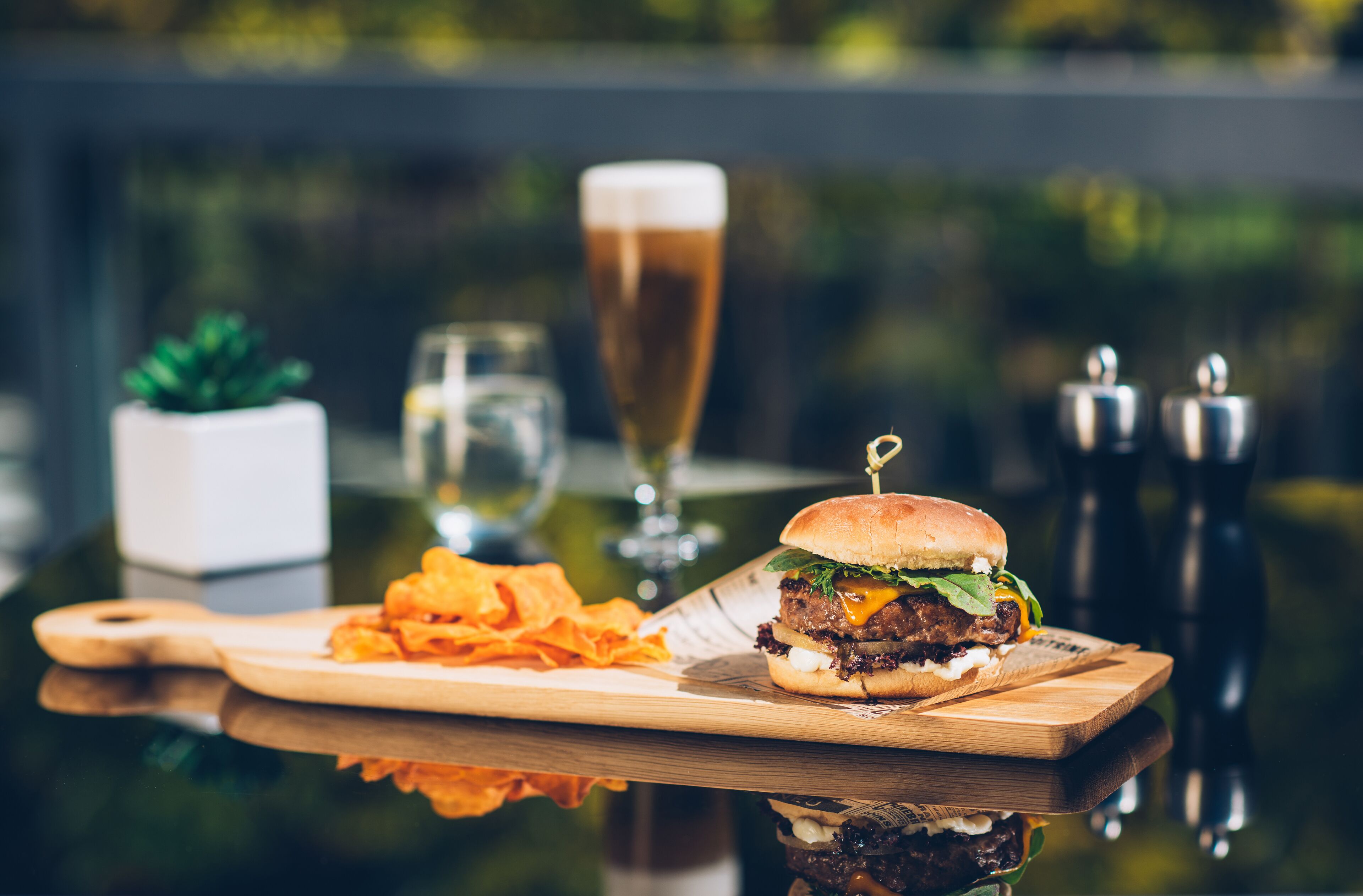 A burger with a side of chips on a wooden board, a glass of beer, and a small potted plant.