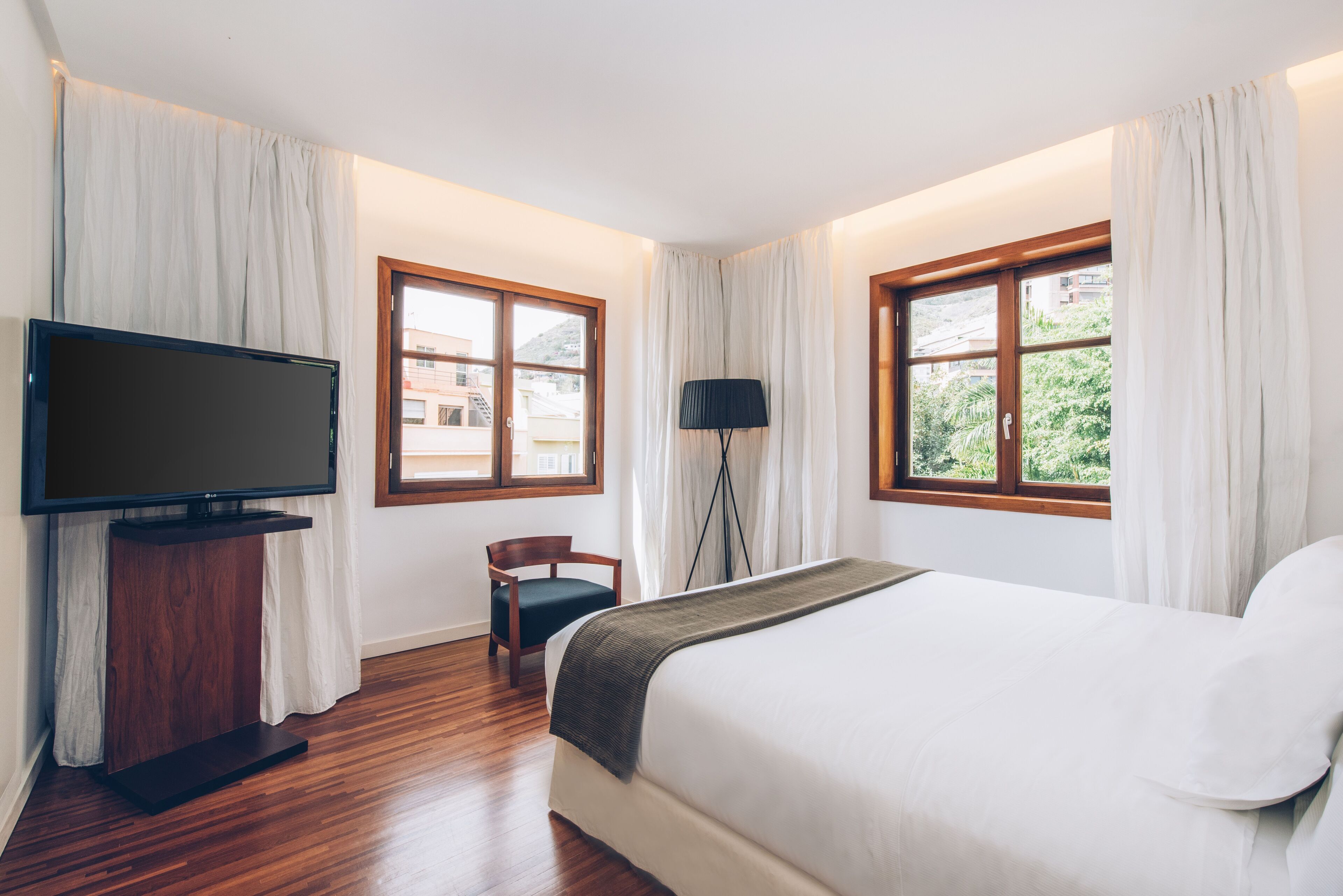 Ein Hotelzimmer mit einem großen Bett, einem Fernseher auf einem Tisch, einem Sessel und zwei mit Vorhängen versehenen Fenstern.