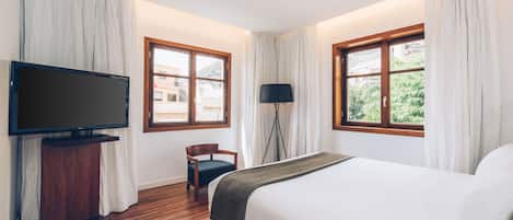 Habitación de hotel con una cama grande, un televisor en un mueble bajo, una silla y dos ventanas con cortinas.