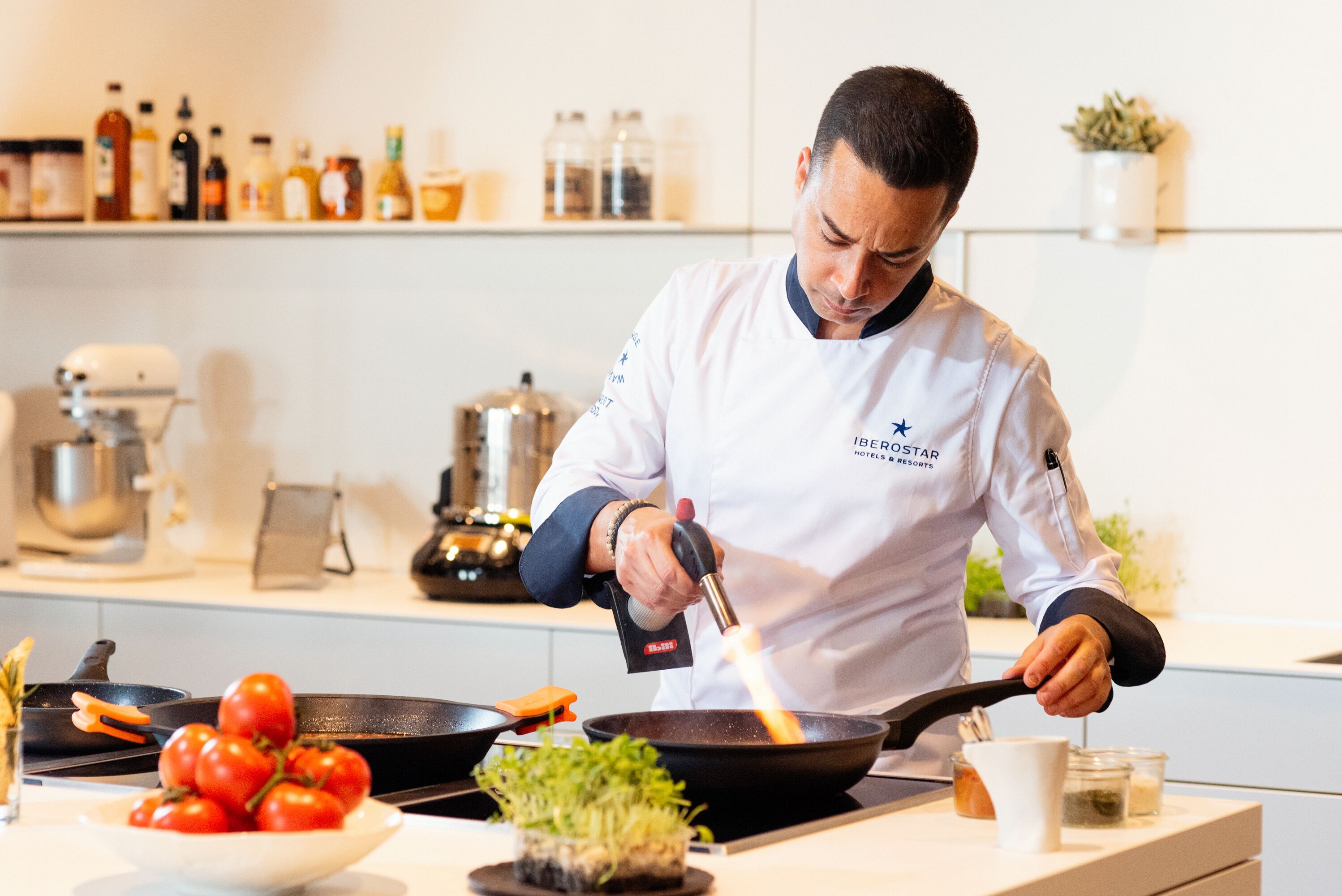 Un chef, vêtu d’un uniforme blanc, cuisine à l’aide d’une poêle sur une cuisinière.