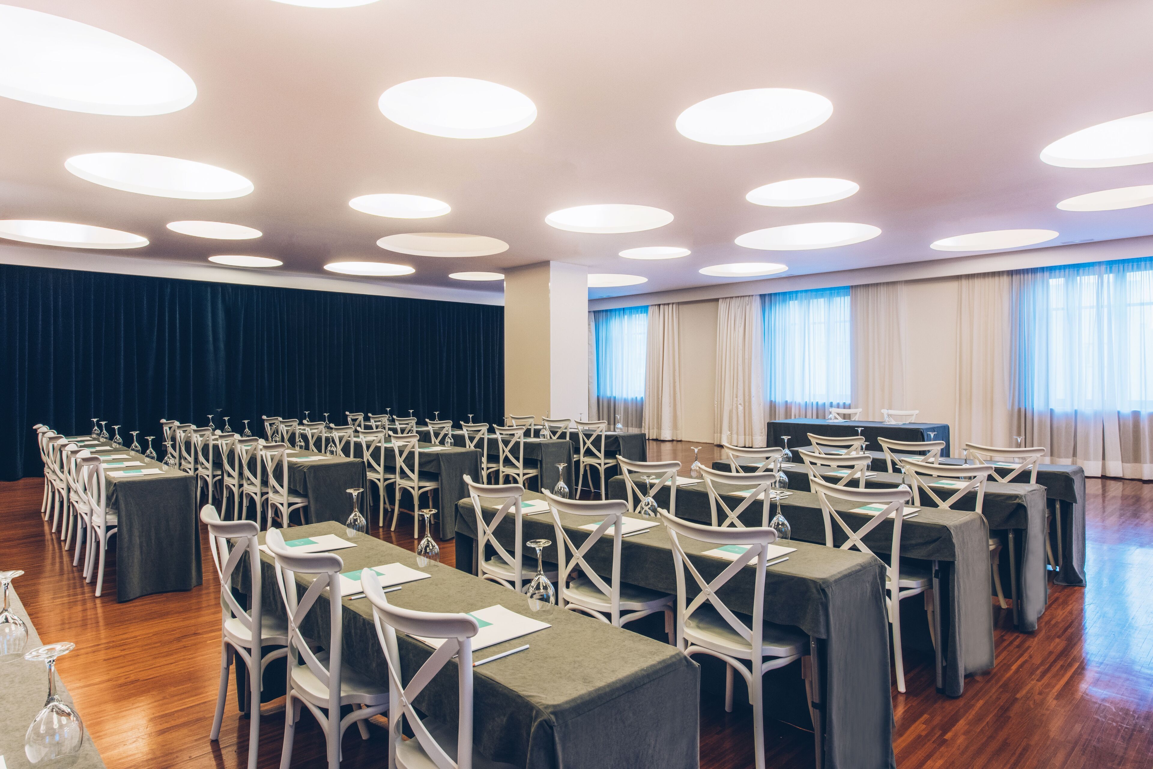 A conference room with rows of tables and chairs, a stage, and large windows with curtains.