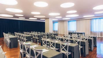 A conference room with rows of tables and chairs, a stage, and large windows with curtains.