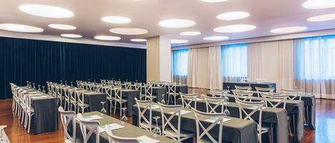 A conference room with rows of tables and chairs, a stage, and large windows with curtains.