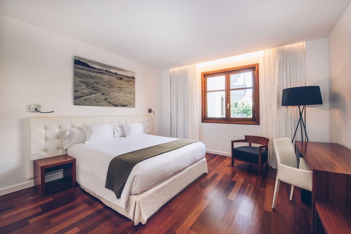 A hotel room with a large bed, a desk, a chair, a lamp, and a window with curtains.