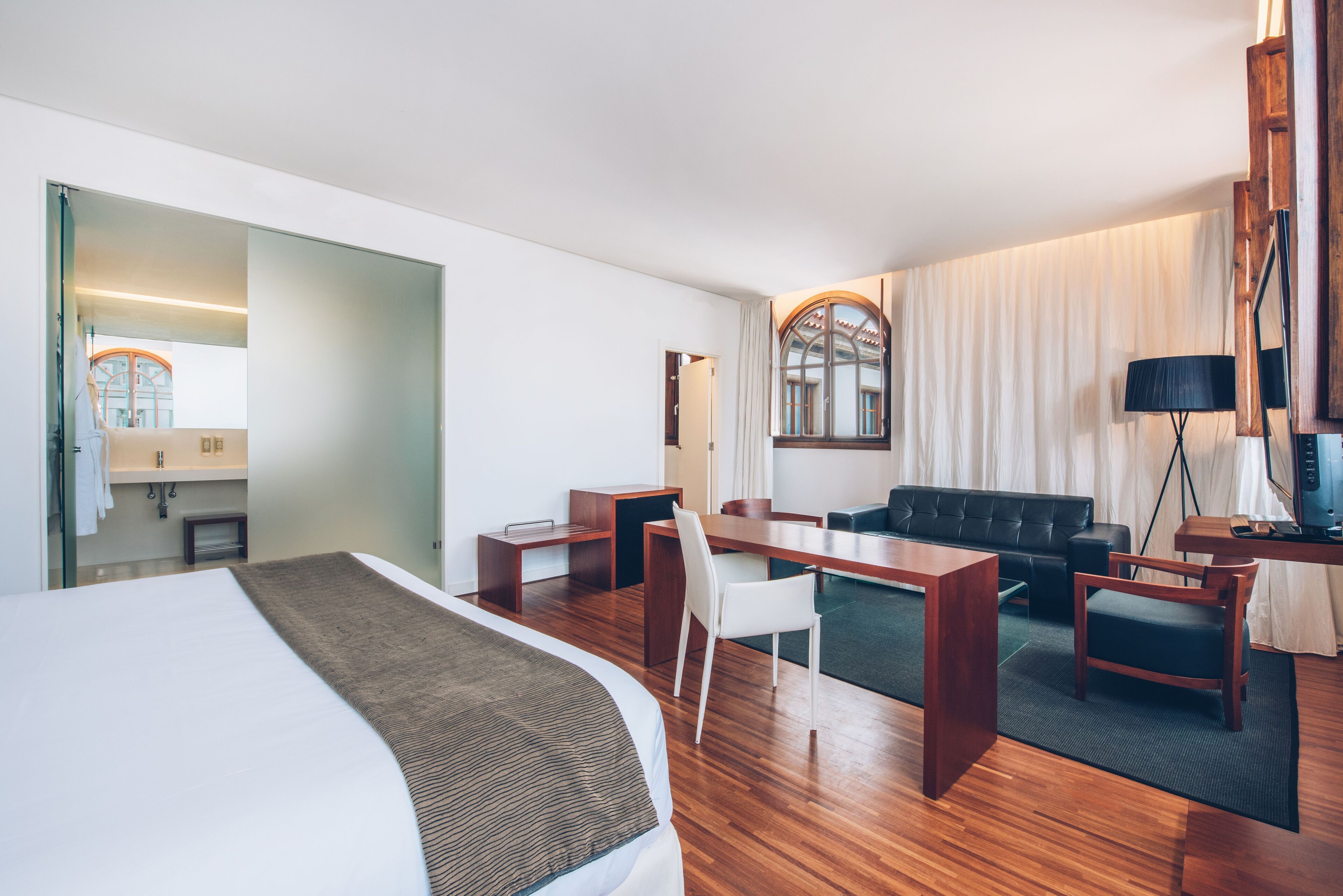 A modern hotel room with a large bed, a desk, a sofa, and a dining table. There is a window with curtains and a mirror on the wall.