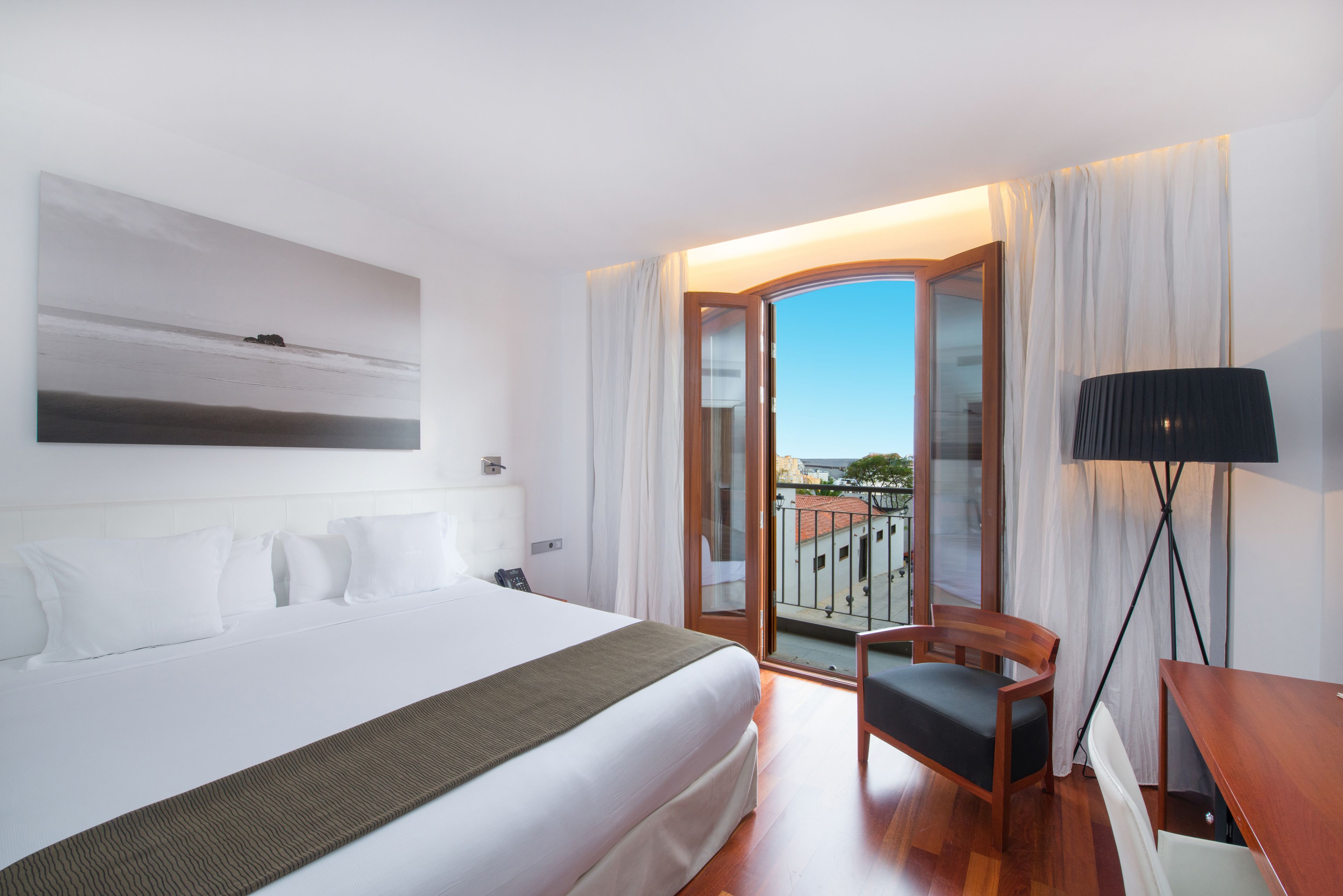 Habitación de hotel con una cama grande, una silla, un escritorio y vistas de la ciudad a través de un ventanal.