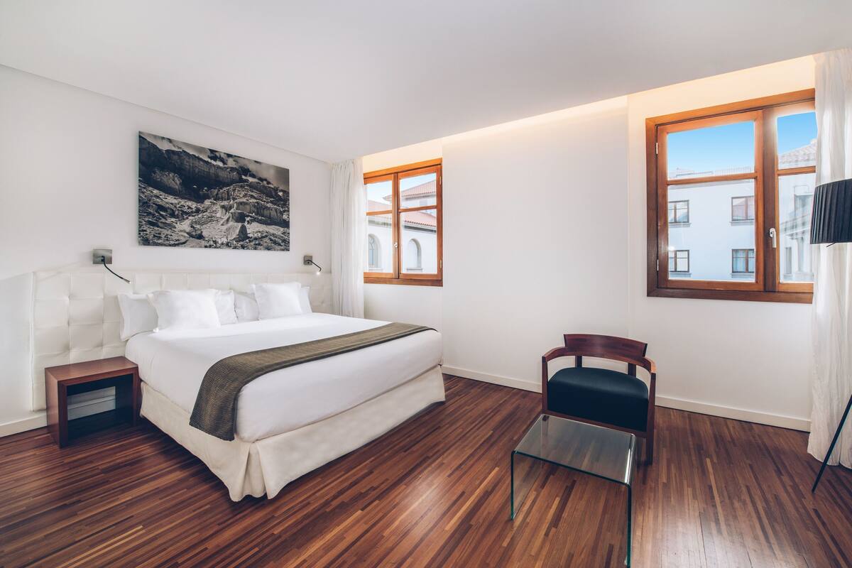 A modern hotel room with a large bed, a wooden floor, a small table, and a chair.