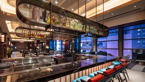 A modern bar area with a long counter, seating with blue and red cushions, and a large window offering a view of the cityscape. - Tempo by Hilton New York Times Square (New York)