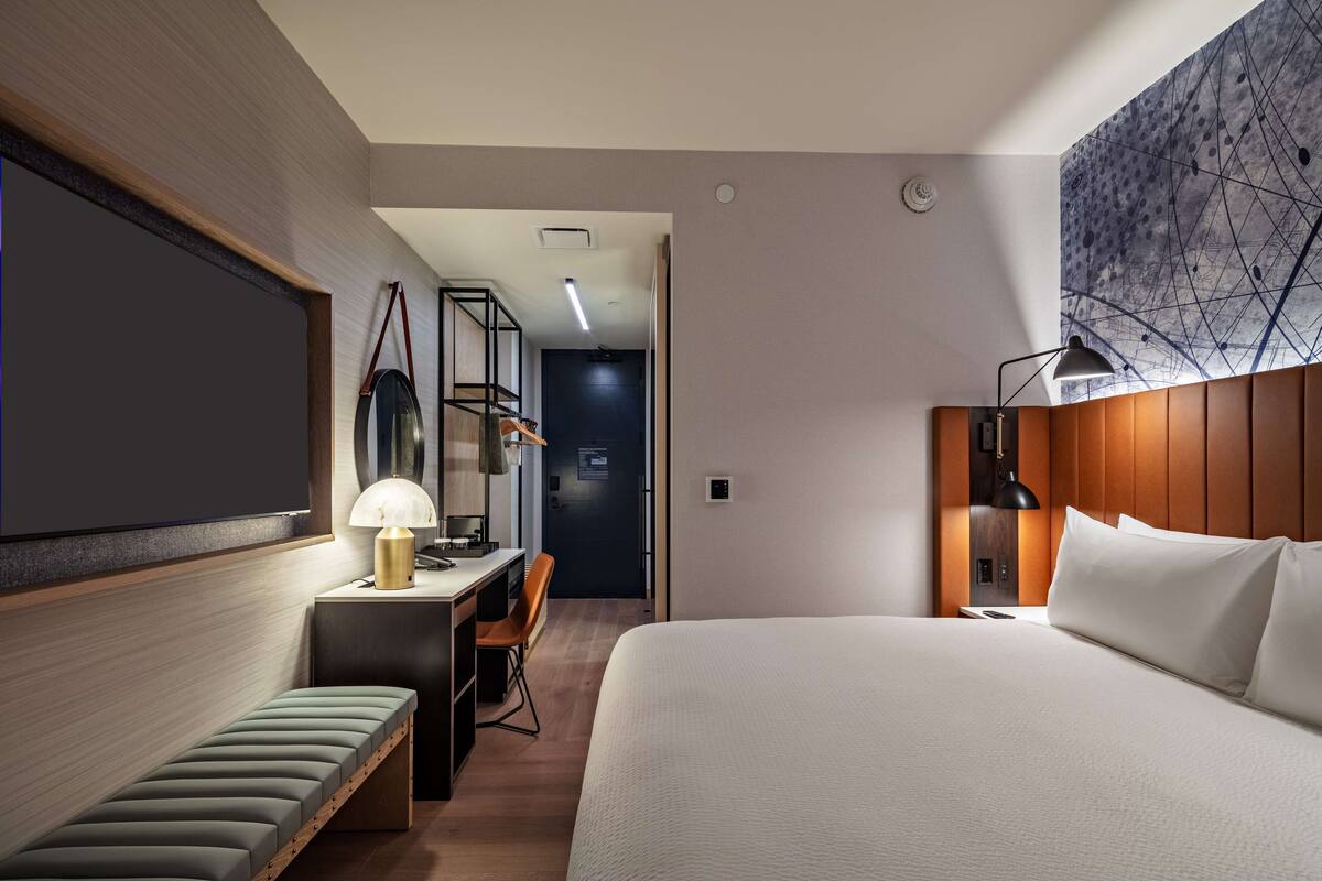 A modern hotel room with a large bed, a desk with a lamp, a bench, and a view of the hallway.