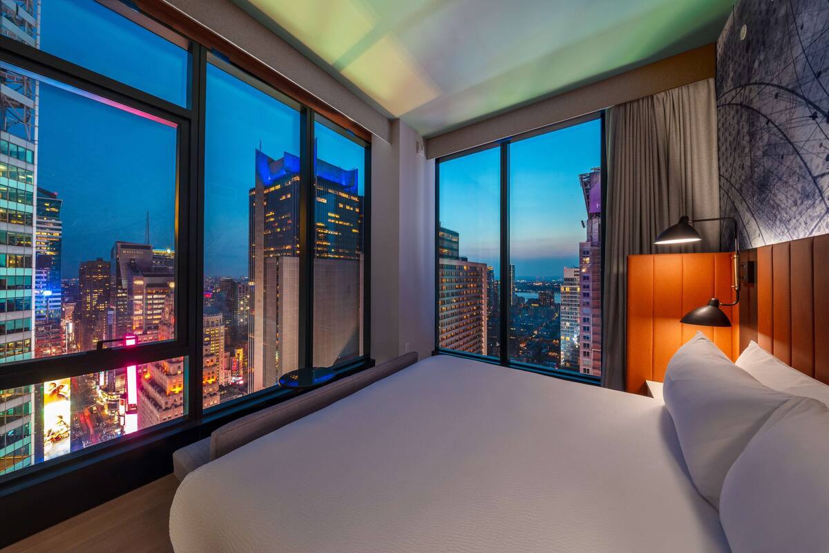 A hotel room with large windows offering a city view at dusk, a bed with white linens, and a modern lamp.