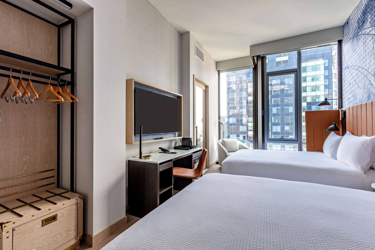 A hotel room with two beds, a desk with a flat-screen TV, a wardrobe, and a window with a city view.
