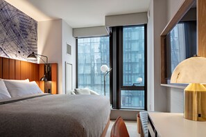 A modern hotel room with a large bed, a desk, and a view of the city through large windows.