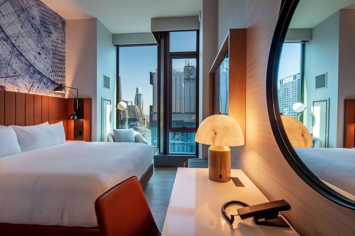 A hotel room with a large bed, a desk with a lamp, a chair, and a view of the city through large windows.