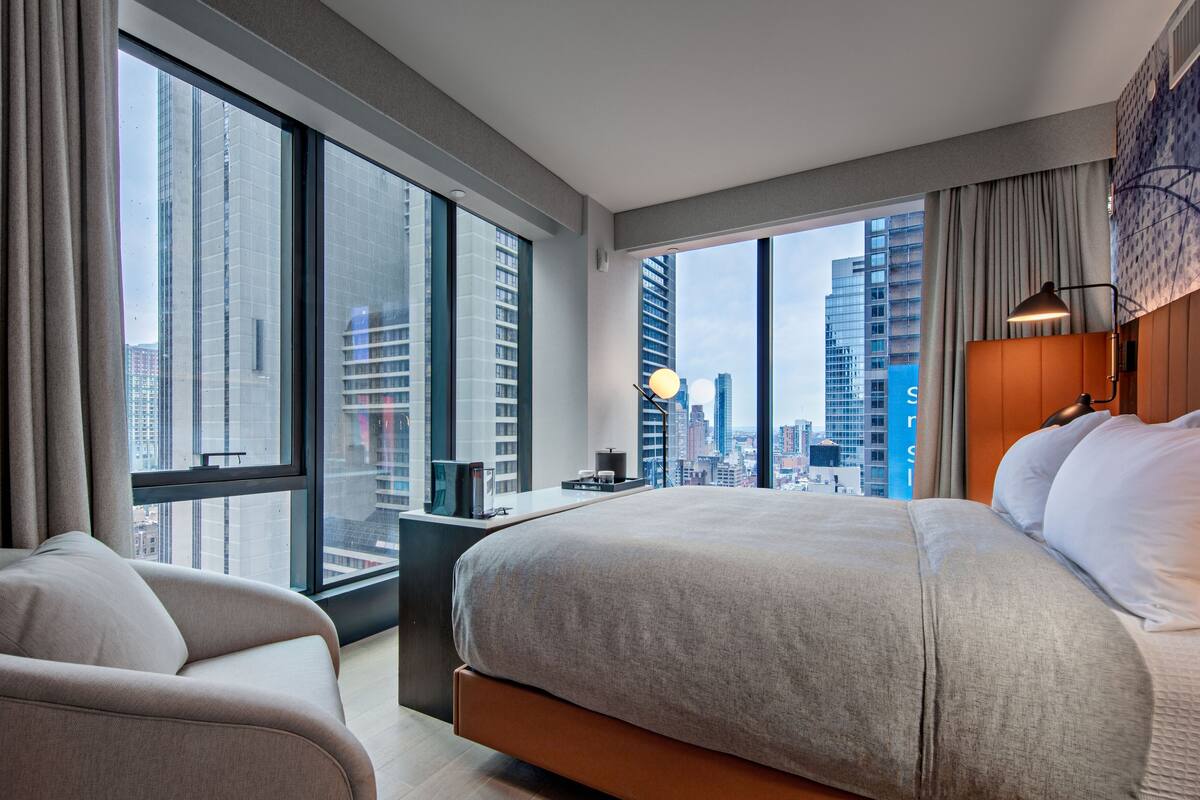 A modern hotel room with a large bed, a desk, and a view of the cityscape through large windows.