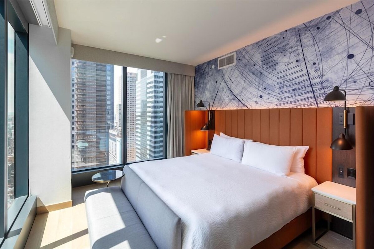 Une chambre d’hôtel moderne, dotée d’un grand lit, d’un bureau et offrant une vue sur les bâtiments de la ville.