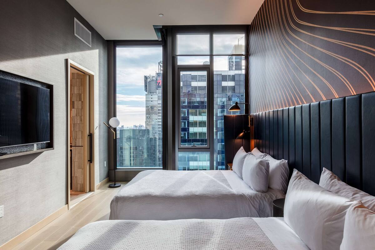 A modern hotel room with a large bed, a TV mounted on the wall, and a city view through large windows.