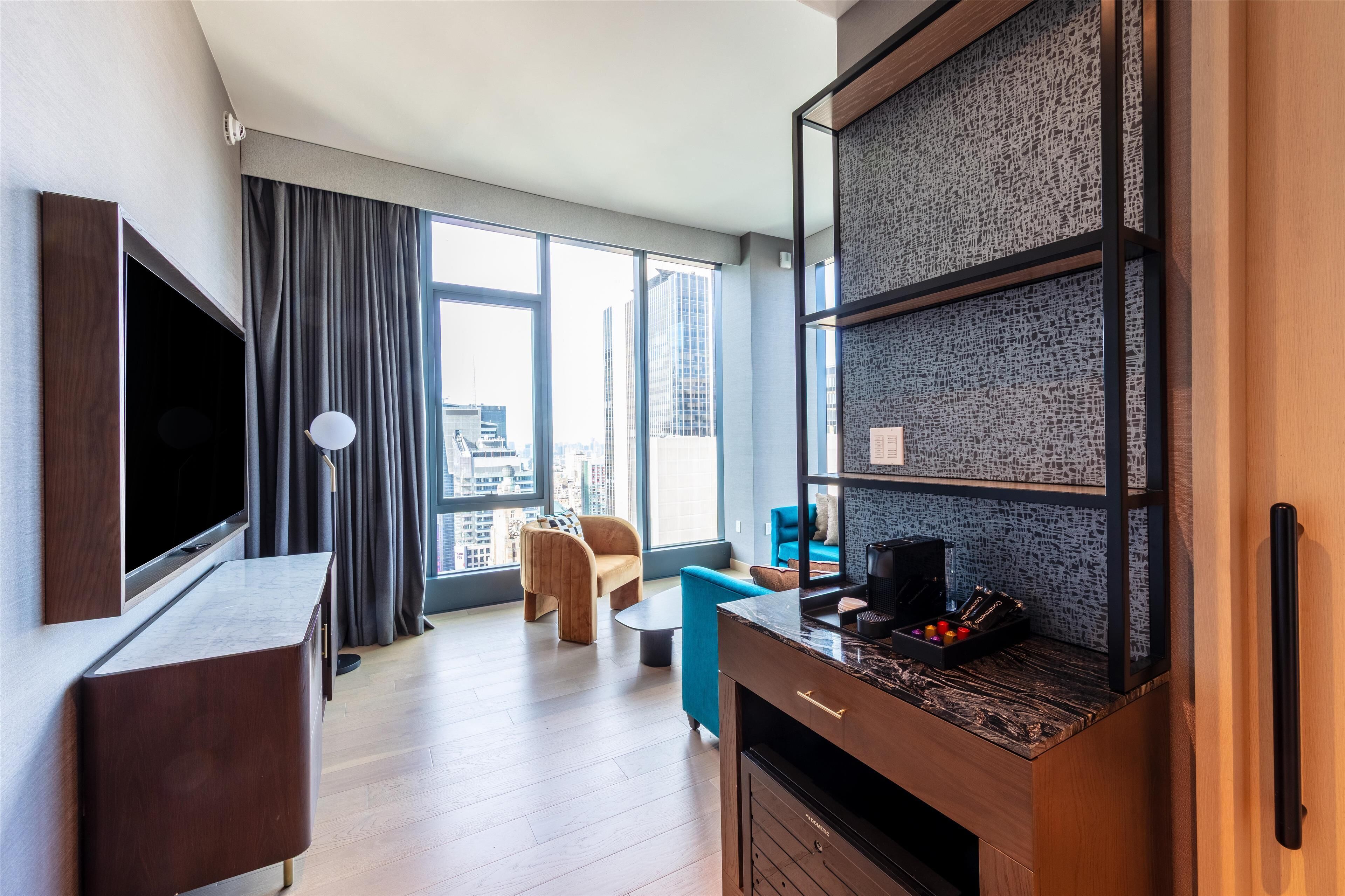 A modern hotel room with a large TV, a dining area with a blue chair, a kitchen area with a marble countertop, and large windows offering a city view.