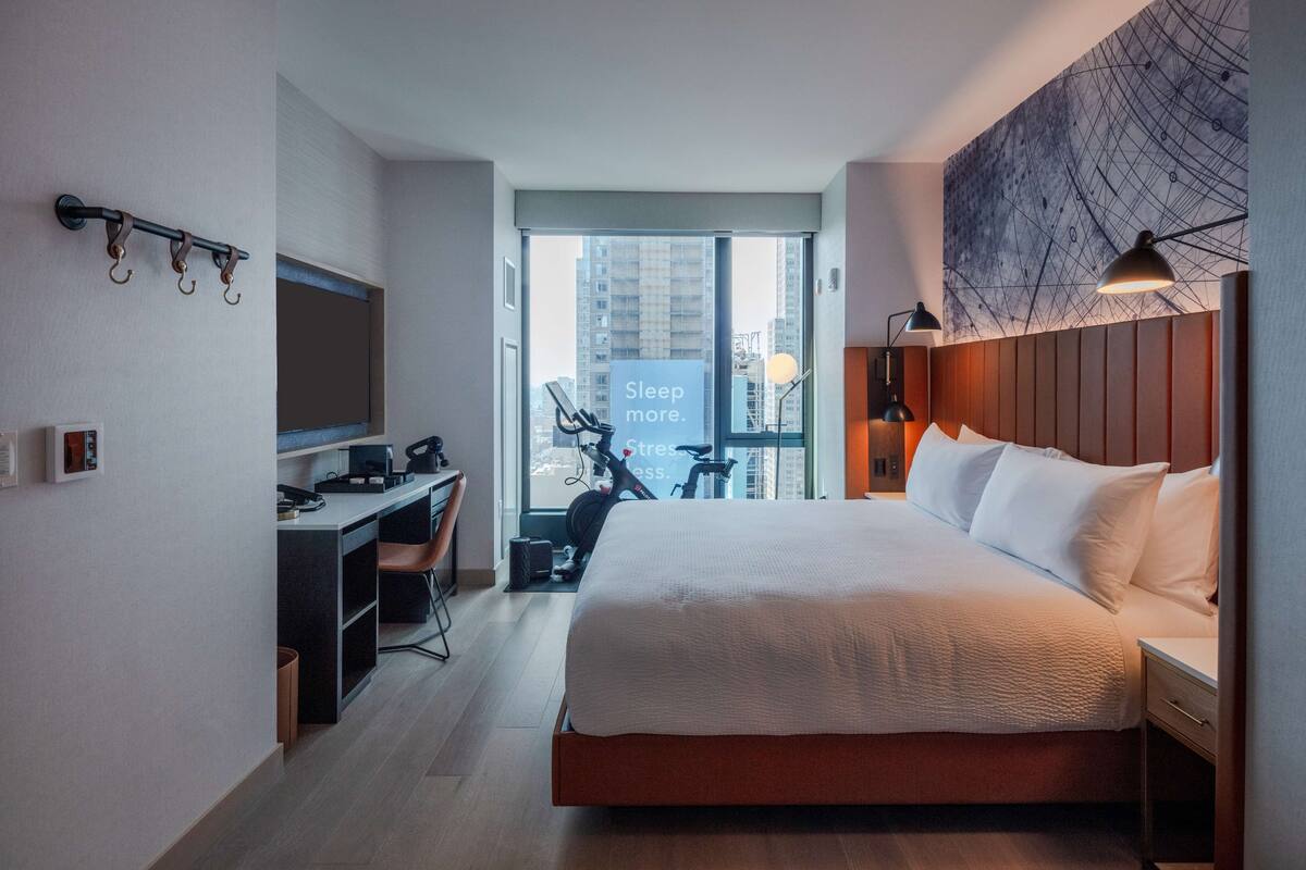 A modern hotel room with a large bed, a desk with a computer, and a view of the city through a large window.