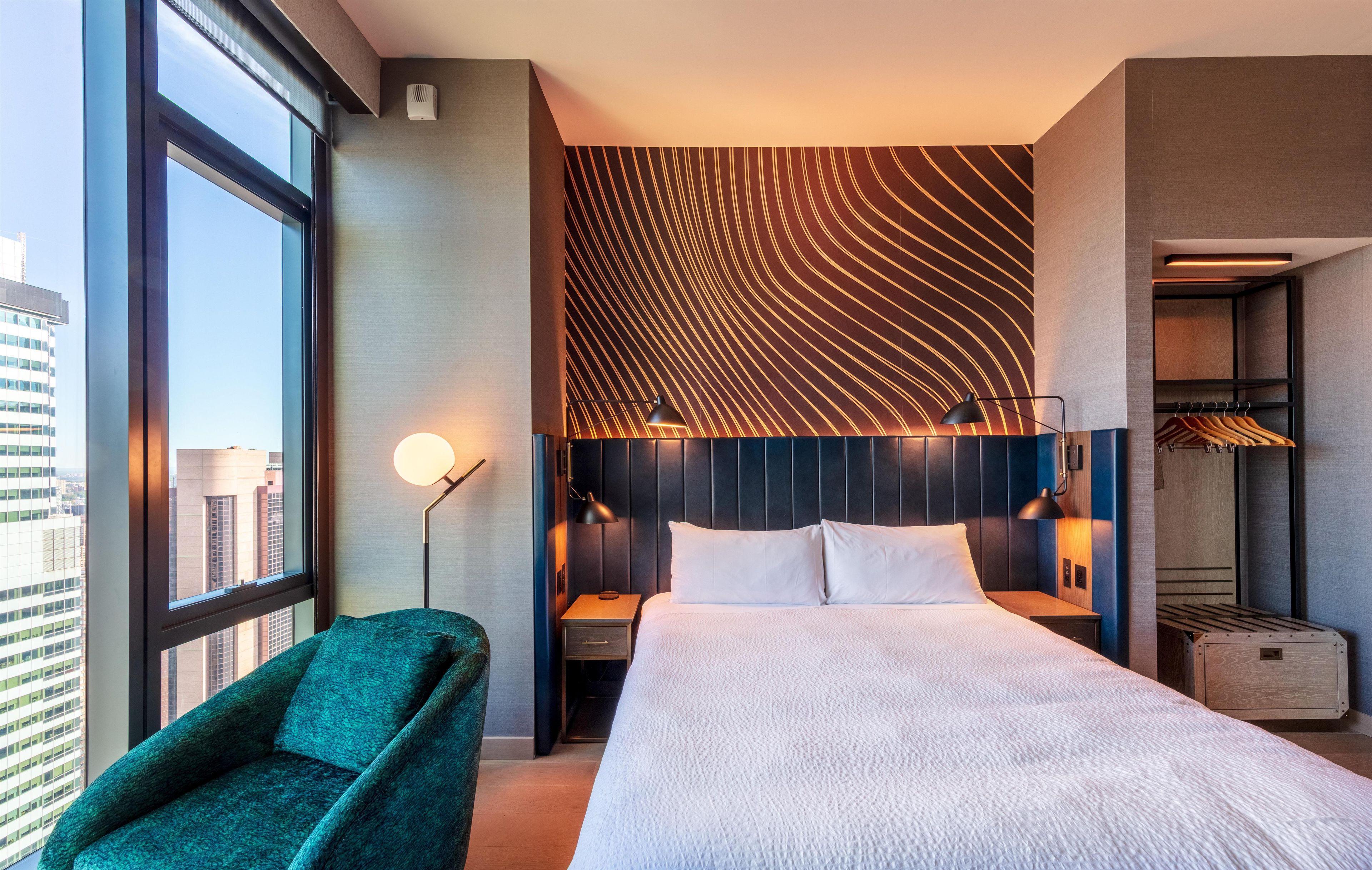 A modern hotel room with a large bed, a seating area, a wardrobe, and a view of the cityscape.