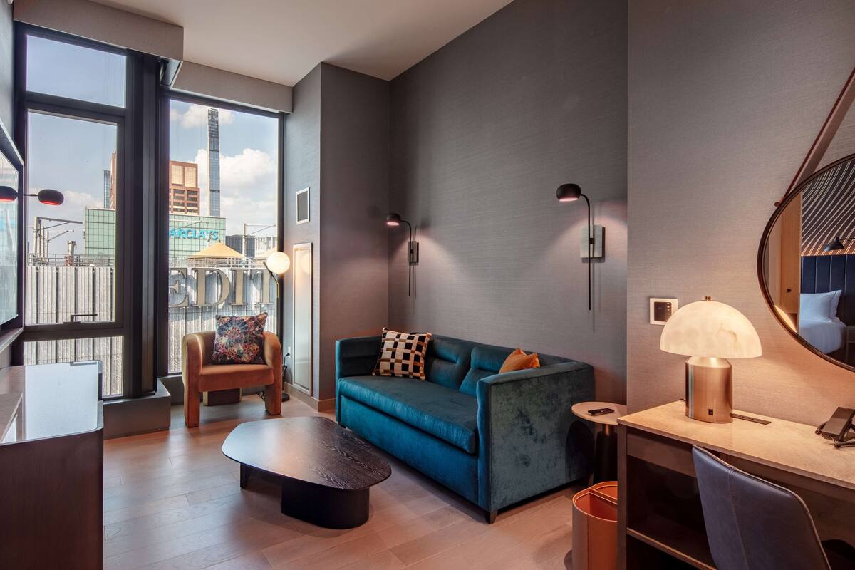 A modern hotel room with a large window, a sofa, a coffee table, a desk, and a mirror.