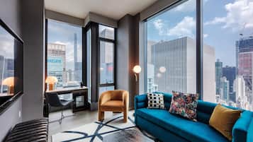 Habitación de hotel moderna con un ventanal grande que ofrece vista a la ciudad, un sofá con cojines decorativos, un escritorio con lámpara y una alfombra estampada.