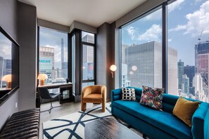 Ein modernes Hotelzimmer mit großem Fenster und Blick auf die Stadt, einer Couch mit dekorativen Kissen, einem Schreibtisch mit Lampe und einem gemusterten Teppich.